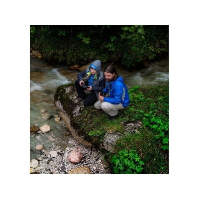 Dve osoby v outdoorovom oblečení, sediace na kameni pri potoku v lese, držia ďalekohľad Steiner Skyhawk 3.0 10x32, ideálne na turistiku a pozorovanie prírody.