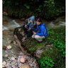 Dve osoby v outdoorovom oblečení, sediace na kameni pri potoku v lese, držia ďalekohľad Steiner Skyhawk 3.0 10x32, ideálne na turistiku a pozorovanie prírody.